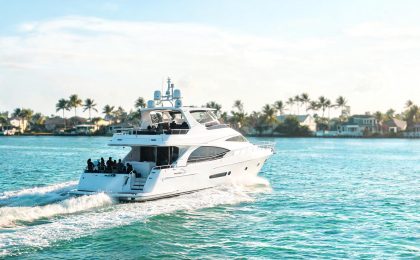 A sleek motoryacht underway on calm turquoise water near Fort Lauderdale with a group of guests enjoying the aft deck, palm-lined Fort Lauderdale coastline visible in the background