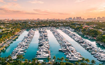 Aerial view of a major Fort Lauderdale marina showing rows of yachts of varying sizes in well-maintained wet slips, with Broward County urban skyline visible in background